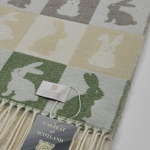 May include: A rectangular scarf with a repeating pattern of white bunny silhouettes on a checkered background of green, beige, and gray squares. The scarf has a fringe and a tag that reads "CALZEAT of SCOTLAND LUXURY SCARF with CASHMERE."