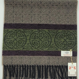 Puede incluir: Bufanda rectangular con diseño de nudo celta. La bufanda presenta un diseño de bloques de color con tonos de marrón, verde y morado. Tiene flecos en la parte inferior y una etiqueta que dice "CALZEAT".