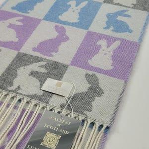 May include: A rectangular scarf with a checkered pattern of bunnies in blue, purple, and gray on a light gray background. The scarf has a fringe and a tag that reads "Luxury Scarf with Cashmere".