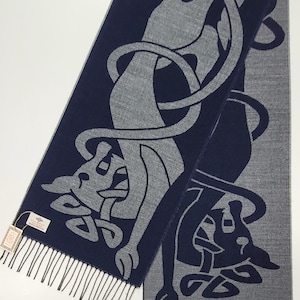May include: A navy and grey scarf featuring a Celtic knot design. The scarf has a fringed edge and a repeating pattern of stylized animal figures and interwoven knotwork. The scarf is made of a soft material.