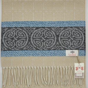 May include: A beige scarf with a blue and gray stripe featuring a Celtic knot design. The scarf has fringe on the bottom and a label that reads "Calzeat -  Irish Collection".