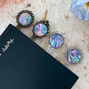 May include: Four ornate metal bookmarks with iridescent abalone shell centers rest on a closed book. The bookmarks have decorative frames and ruler-like stems. The book is dark blue, and the background is a textured, light-colored surface with floral accents.