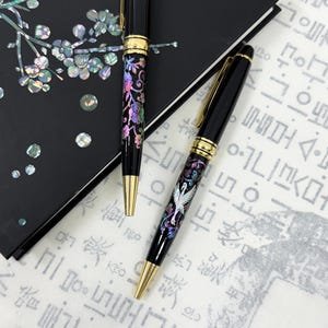 Pode incluir: Duas canetas pretas com detalhes dourados e intrincados desenhos florais e de pássaros. As canetas têm um acabamento brilhante e estão colocadas perto de um caderno preto com detalhes em madrepérola. As pontas das canetas são douradas.