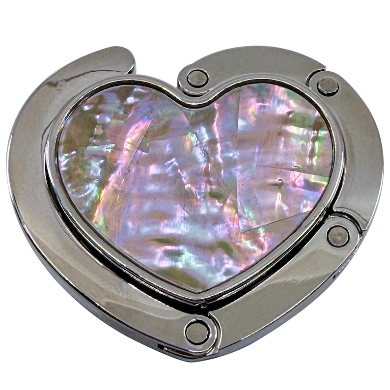 K&ouml;nnte beinhalten: Ein herzf&ouml;rmiger Handtaschenhaken mit polierter Silberoberfl&auml;che. Die Mitte zeigt ein schimmerndes, irisierendes Abalone-Muscheldesign. Der Haken ist zum Aufh&auml;ngen einer Handtasche an einer Tisch- oder Thekenkante konzipiert.
