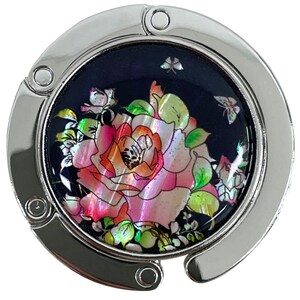 May include: A silver-toned purse hook with a decorative, circular center. The center features a dark blue background with a large, pink and orange rose and green leaves. Two small butterflies are also visible.