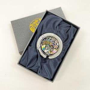May include: A silver-toned purse hook with a colorful peacock design, presented in a gray gift box with a gold emblem. The hook features a detailed peacock with blue, yellow, and pink accents, set against a black background.