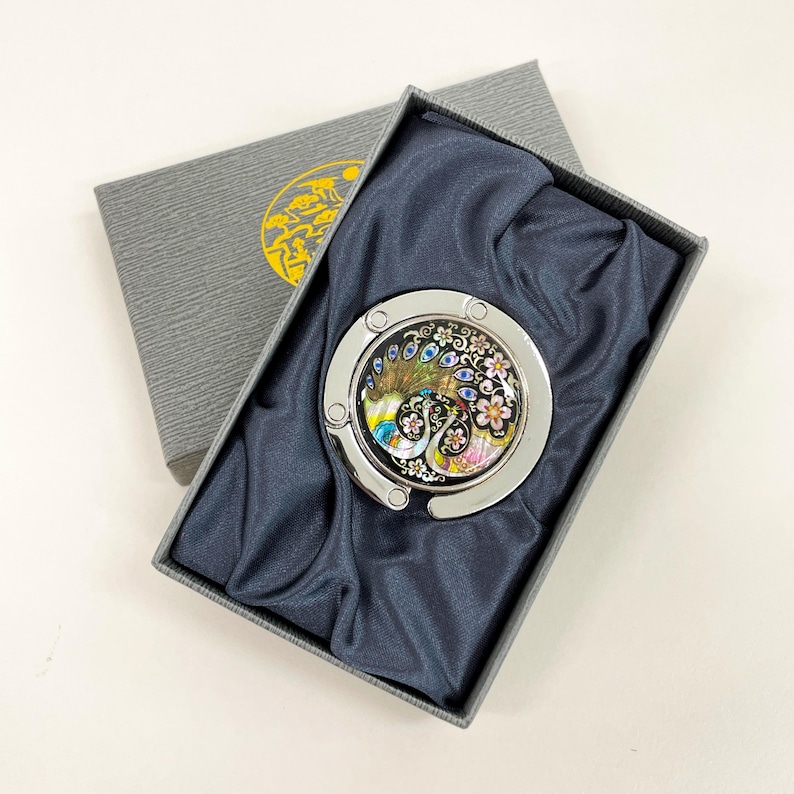 May include: A silver-toned purse hook with a colorful peacock design, presented in a gray gift box with a gold emblem. The hook features a detailed peacock with blue, yellow, and pink accents, set against a black background.