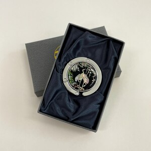May include: A silver-toned purse hook with a black and iridescent crane design, presented in a gray gift box with a dark blue satin lining. The crane is depicted in a naturalistic setting.