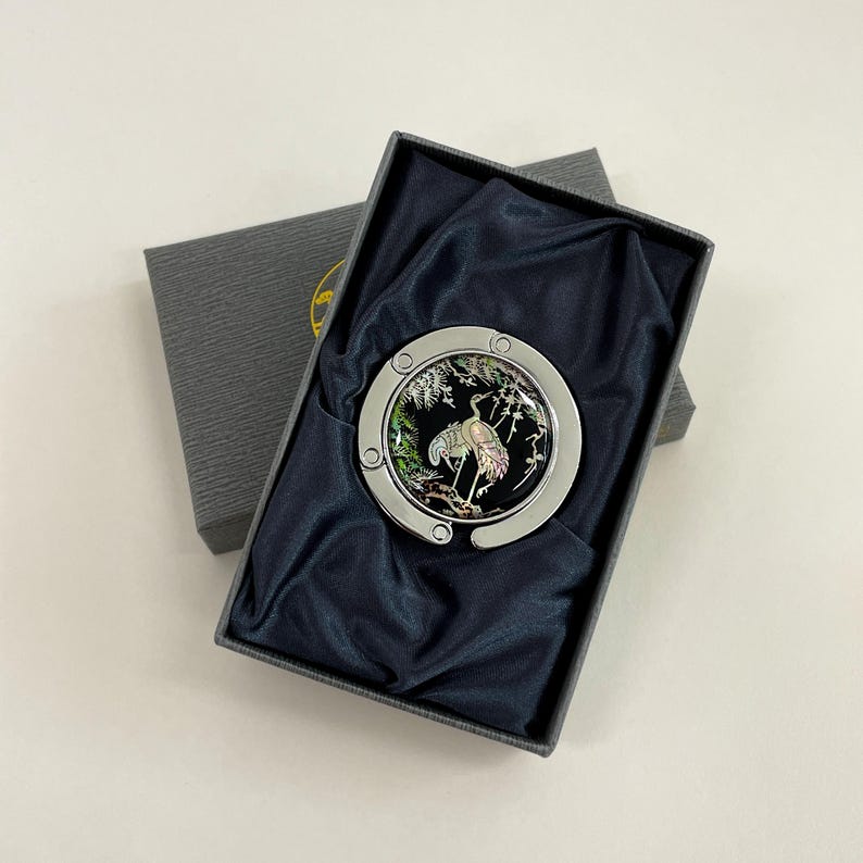 May include: A silver-toned purse hook with a black and iridescent crane design, presented in a gray gift box with a dark blue satin lining. The crane is depicted in a naturalistic setting.