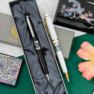 May include: Two stylish pens, one black and one white, displayed in a grey box with a satin lining. The black pen has a geometric design with iridescent details. The white pen features gold-coloured trim and a similar iridescent pattern. The pens are on a green surface.