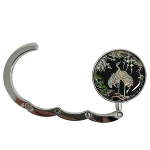 May include: A silver-toned purse hook with a circular, black decorative element. The circular element features a crane design with iridescent accents. The hook is designed to hang a purse from a table or other surface.