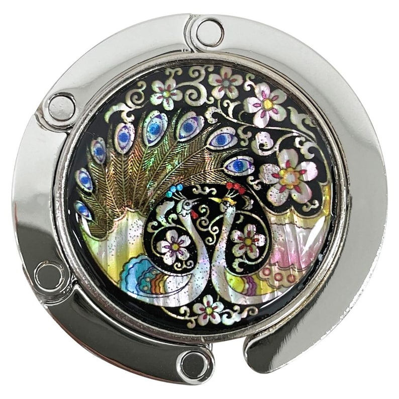 May include: A silver-toned purse hook with a decorative, round centerpiece. The centerpiece features a black background with a peacock and floral design in iridescent colors. The hook is designed to hang a purse from a table.
