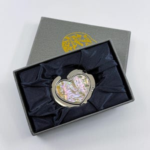 K&ouml;nnte beinhalten: Ein herzf&ouml;rmiger Handtaschenhaken mit Perlmutt-Inlay, pr&auml;sentiert in einer grauen Geschenkbox mit dunkelblauem Satinfutter. Der Haken ist silberfarben und l&auml;sst sich zur Aufbewahrung zusammenklappen. Die Box hat ein goldenes Emblem auf dem Deckel.