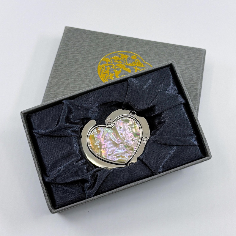 K&ouml;nnte beinhalten: Ein herzf&ouml;rmiger Handtaschenhaken mit Perlmutt-Inlay, pr&auml;sentiert in einer grauen Geschenkbox mit dunkelblauem Satinfutter. Der Haken ist silberfarben und l&auml;sst sich zur Aufbewahrung zusammenklappen. Die Box hat ein goldenes Emblem auf dem Deckel.