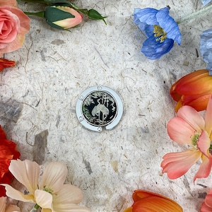 May include: A silver and black purse hook with a crane design, surrounded by colorful flowers. The flowers include red poppies, pink roses, and blue and orange tulips, arranged on a textured, off-white surface.