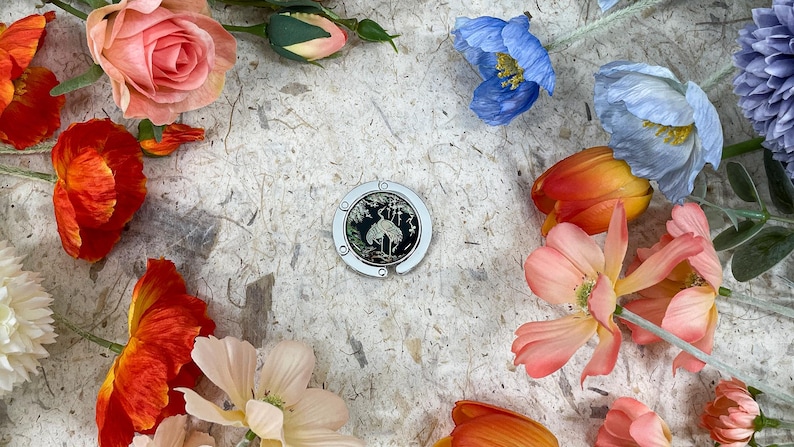 May include: A silver and black purse hook with a crane design, surrounded by colorful flowers. The flowers include red poppies, pink roses, and blue and orange tulips, arranged on a textured, off-white surface.