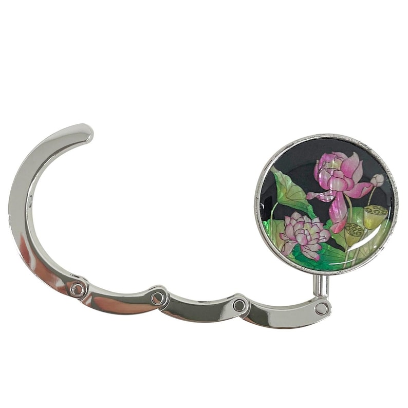 May include: A silver-colored purse hook with a circular, black decorative element. The decorative element features a pink lotus flower design with green leaves and yellow seed pods. The hook is designed to hang a purse from a table or other surface.