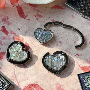 K&ouml;nnte beinhalten: Drei herzf&ouml;rmige Taschenaufh&auml;nger mit irisierenden Abalone-Muscheleins&auml;tzen und schwarzen Metallrahmen. Ein Aufh&auml;nger ist offen und zeigt den Haken. Die anderen beiden sind geschlossen. Der Hintergrund ist eine rosa-rot gemusterte Oberfl&auml;che.