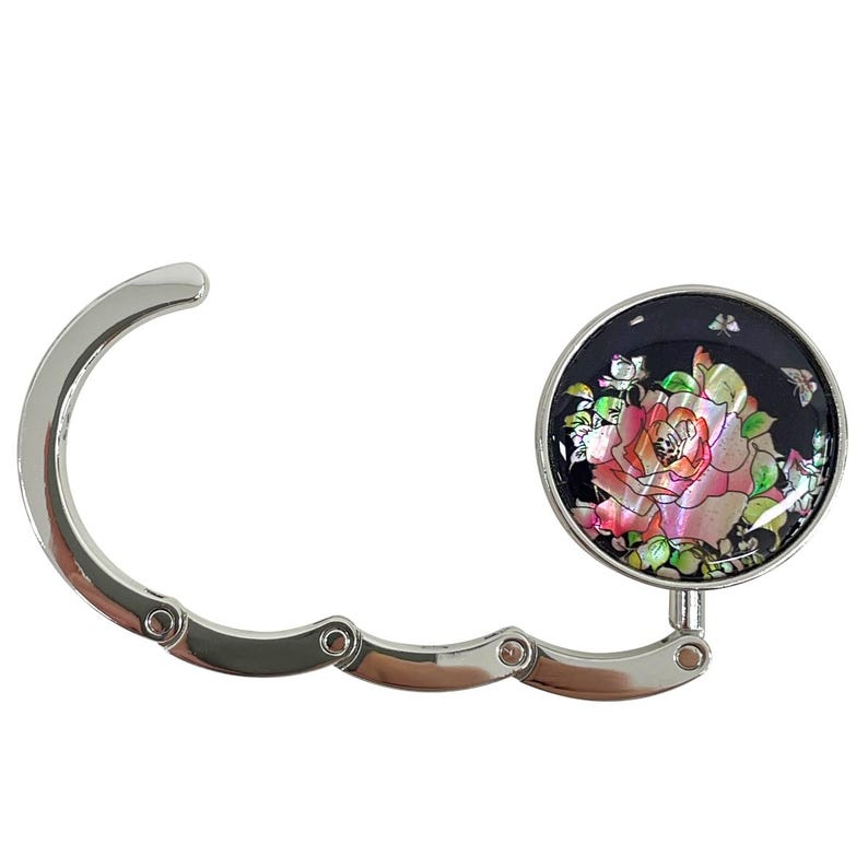 May include: A silver purse hook with a decorative round top. The top features a pink and iridescent rose design on a black background. The hook is designed to hang a purse from a table or counter edge.