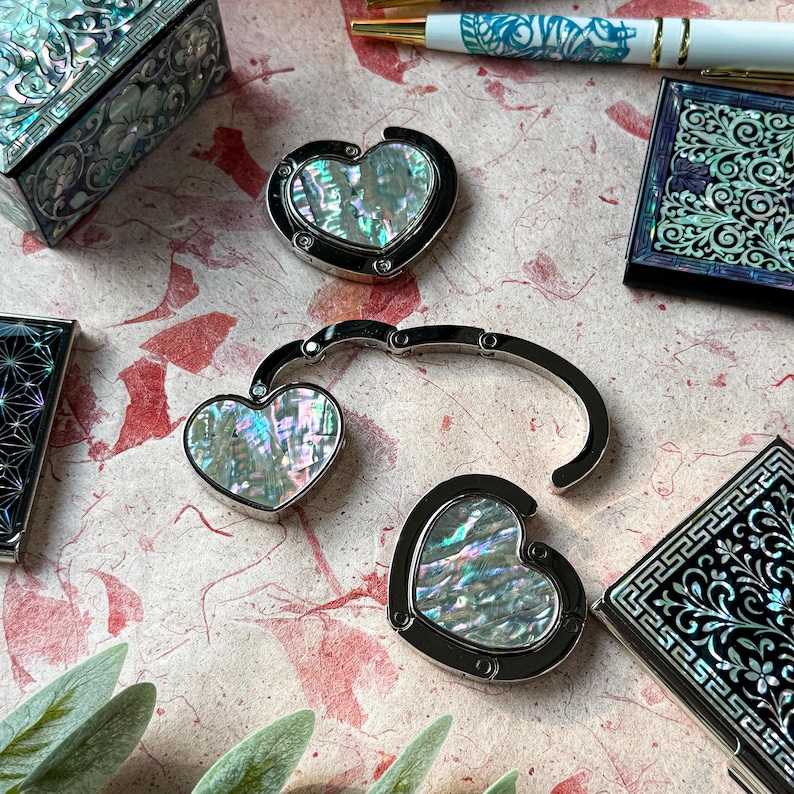 K&ouml;nnte beinhalten: Eine Sammlung von schillernden herzf&ouml;rmigen Taschenhaltern und dekorativen Boxen. Die herzf&ouml;rmigen Aufh&auml;nger haben einen silbernen Rahmen und eine Perlmutt-Einlage. Die Boxen weisen aufwendige Designs auf. Stifte mit blauen und goldenen Akzenten sind ebenfalls sichtbar.