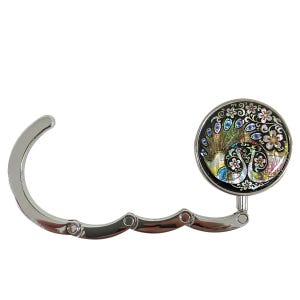 May include: A silver-colored purse hook with a decorative round top. The top features a colorful peacock design with blue, green, and gold accents, set against a black background. The hook is designed to fold for storage.