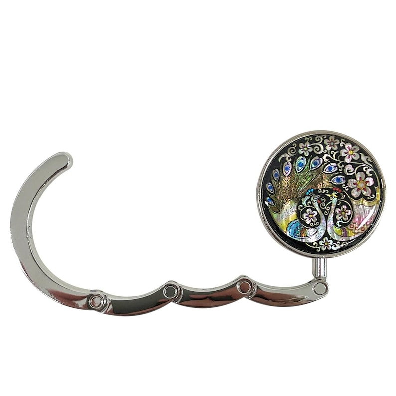 May include: A silver-colored purse hook with a decorative round top. The top features a colorful peacock design with blue, green, and gold accents, set against a black background. The hook is designed to fold for storage.