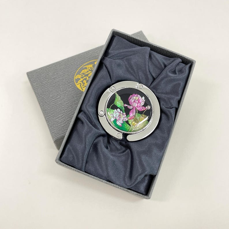 May include: A silver-toned purse hook with a floral design, presented in a gray gift box. The hook features a black background with pink and white flowers and green leaves. The box is lined with dark gray fabric.