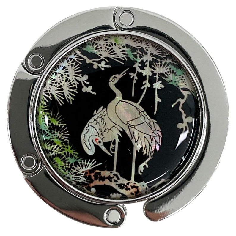 May include: A silver-toned purse hook with a round, decorative center. The center features a black background with a mother-of-pearl crane design, set against a backdrop of stylized branches and foliage. The hook is designed to hang from a table.