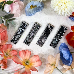 May include: Four rectangular money clips with decorative black and white floral and nature designs. The clips are surrounded by colourful flowers, including pink, peach, and blue blooms, creating a visually appealing arrangement.