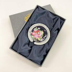May include: A silver-toned purse hook with a black circular face featuring a pink and red rose design. The hook is presented in a gray gift box with a dark blue satin lining, ideal for gifting.