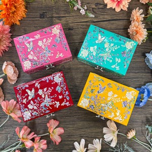 May include: Four rectangular jewelry boxes in pink, teal, red, and yellow. Each box features a butterfly and floral design with silver accents. The boxes have a silver clasp and are arranged on a wooden surface.