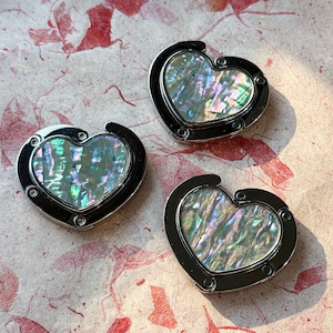 Pode incluir: Três cabides de bolsa em forma de coração com inserções de concha de abalone iridescente e armações de metal prateado. Os cabides são exibidos em uma superfície texturizada rosa e vermelha. Cada cabide tem um design articulado.