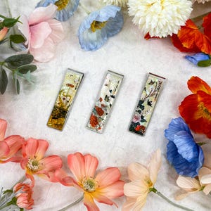 Könnte beinhalten: Drei rechteckige Geldklammern aus Metall mit Blumen- und Schmetterlingsmotiven. Die Klammern weisen eine Vielzahl von Farben auf, darunter Gold, Rot und Grün, und sind von bunten Kunstblumen umgeben. Die Klammern sind etwa 7,6 cm lang.