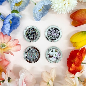 Könnte beinhalten: Vier silberfarbene Handtaschenaufhänger mit dekorativen Oberteilen mit Schmetterlings- und Blumendesigns. Die Designs umfassen schwarze, weiße und farbenfrohe Akzente. Die Aufhänger werden auf einer rosa Oberfläche präsentiert, umgeben von Blumen.