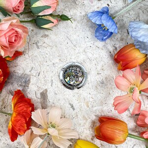 May include: A silver purse hook with an abalone shell design is surrounded by colorful artificial flowers, including roses, tulips, and poppies. The flowers are arranged on a textured, off-white surface, creating a vibrant and decorative display.