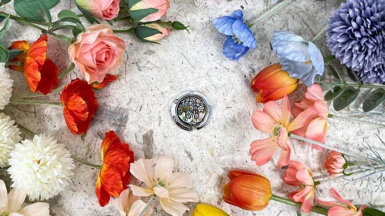 May include: A silver purse hook with an abalone shell design is surrounded by colorful artificial flowers, including roses, tulips, and poppies. The flowers are arranged on a textured, off-white surface, creating a vibrant and decorative display.