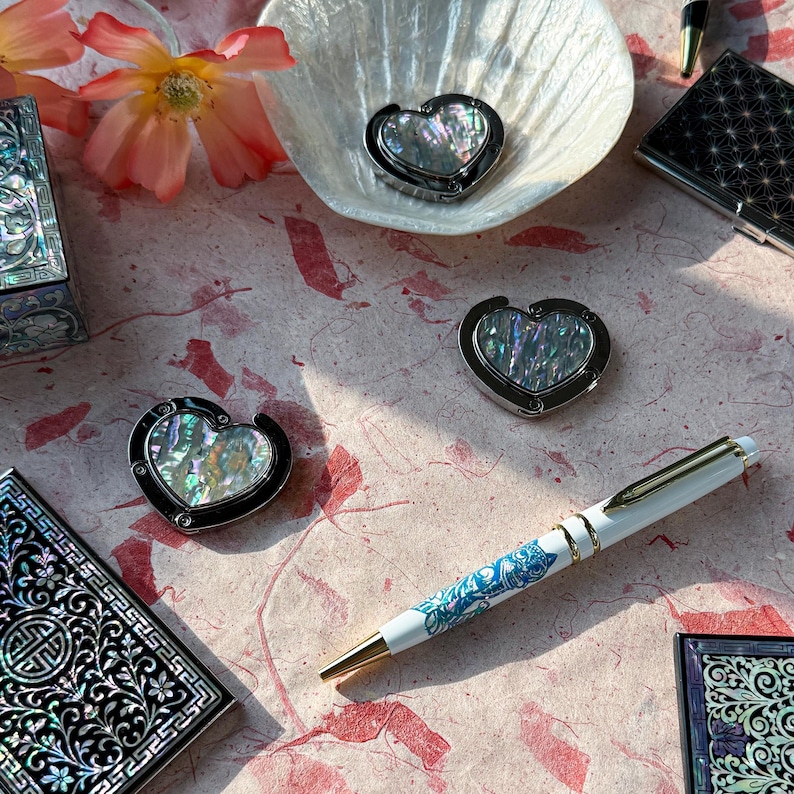K&ouml;nnte beinhalten: Eine Auswahl an Dekorationsartikeln, darunter herzf&ouml;rmige Taschenaufh&auml;nger mit schillernden Einlagen, ein wei&szlig;er Stift mit blauen Blumenmustern und kleine, verzierte Schachteln. Die Artikel sind auf einer rosa gemusterten Oberfl&auml;che angeordnet.