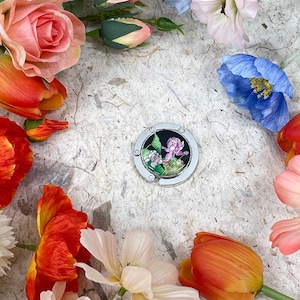 May include: A silver purse hook with a floral design, surrounded by colorful flowers. The flowers include orange tulips, pink roses, white dahlias, and blue poppies, creating a vibrant and artistic composition.