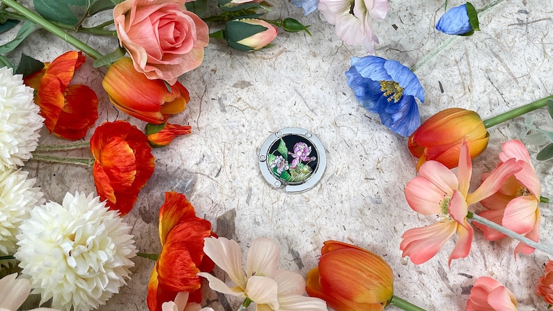 May include: A silver purse hook with a floral design, surrounded by colorful flowers. The flowers include orange tulips, pink roses, white dahlias, and blue poppies, creating a vibrant and artistic composition.