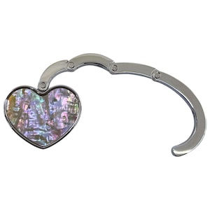 K&ouml;nnte beinhalten: Ein silberfarbener Handtaschenhaken mit herzf&ouml;rmigem Design aus schillerndem Abalone-Muschelmaterial. Der Haken ist klappbar und l&auml;sst sich zusammenfalten. Der herzf&ouml;rmige Teil ist silberfarben umrandet.