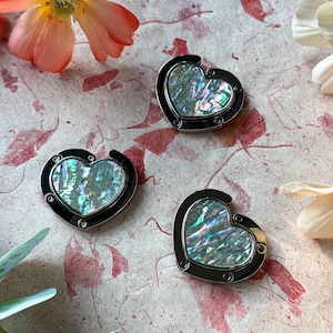 K&ouml;nnte beinhalten: Drei herzf&ouml;rmige Handtaschenaufh&auml;nger mit schillernden Abalone-Muschel-Mittelst&uuml;cken und schwarzen Metallrahmen. Die Aufh&auml;nger werden auf einer strukturierten, gemusterten Oberfl&auml;che mit floralen Akzenten pr&auml;sentiert.