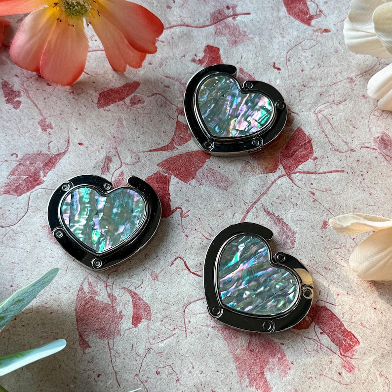 K&ouml;nnte beinhalten: Drei herzf&ouml;rmige Handtaschenaufh&auml;nger mit schillernden Abalone-Muschel-Mittelst&uuml;cken und schwarzen Metallrahmen. Die Aufh&auml;nger werden auf einer strukturierten, gemusterten Oberfl&auml;che mit floralen Akzenten pr&auml;sentiert.
