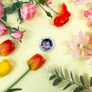 May include: A floral-themed image featuring a silver purse hook with a rose design, surrounded by colorful flowers. The flowers include pink roses, orange and yellow tulips, and other delicate blooms, all arranged on a light yellow background.