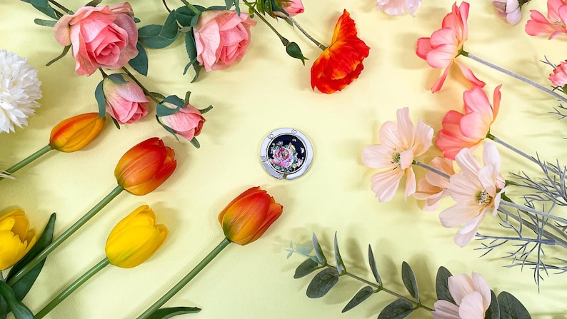 May include: A floral-themed image featuring a silver purse hook with a rose design, surrounded by colorful flowers. The flowers include pink roses, orange and yellow tulips, and other delicate blooms, all arranged on a light yellow background.