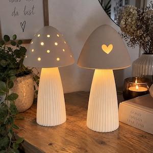 Könnte beinhalten: Zwei weiße pilzförmige Tischlampen mit gerippten Sockeln. Die linke Lampe hat einen Schirm mit kreisförmigen Ausstanzungen, während die rechte Lampe eine herzförmige Ausstanzung aufweist. Beide Lampen strahlen ein warmes, sanftes Licht aus.