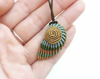 May include: A handmade macrame pendant necklace featuring a seashell design. The pendant has a spiral center in gold, with green, blue, and orange accents. The necklace cord is brown.