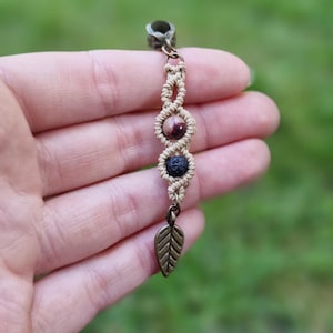 Może przedstawiać: Urok makramy z brązowymi, czerwonymi i czarnymi koralikami, ozdobiony brązowym liściem. Urok zawieszony jest na metalowej pętli, idealny do bransoletki lub naszyjnika.