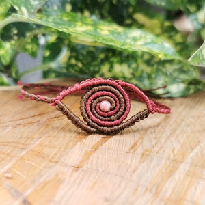 May include: A handmade macrame bracelet featuring a spiral eye design. The bracelet is crafted with brown and pink cord, with a pink bead at the center of the spiral. The bracelet is displayed on a wooden surface.