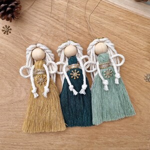 May include: Three handmade angel decorations with wooden heads, white rope hair, and tassel dresses in mustard yellow, teal, and sage green. Each angel has a gold halo and a snowflake. They hang from twine.
