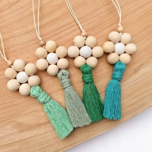 Könnte beinhalten: Vier dekorative Holzperlen-Ornamente mit Quasten. Jedes Ornament hat ein blütenartiges Design aus Naturholzperlen und weißen Perlen. Die Quasten sind in Grün- und Türkistönen gehalten und an einer natürlichen Kordel befestigt.